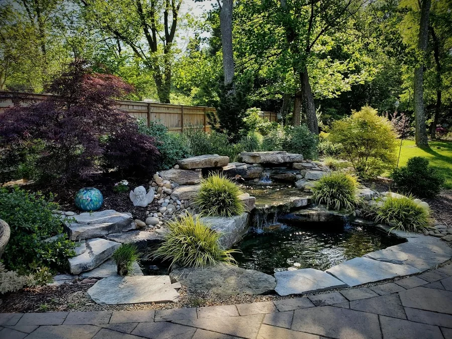 A layered stone waterfall and koi pond built from hand-placed boulders and river rock, tucked into a wooded corner of the yard. Part of a full backyard build in the Upstate.